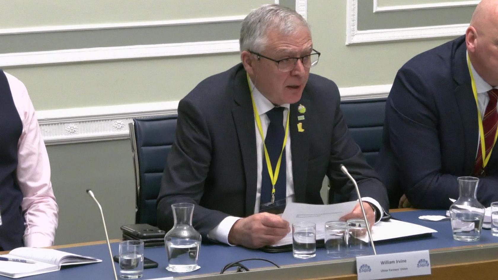 William Irvine at the Stormont committee. He has grey hair, wearing glasses, a dark suit and white shirt. He is holding a piece of paper and speaking into a microphone.