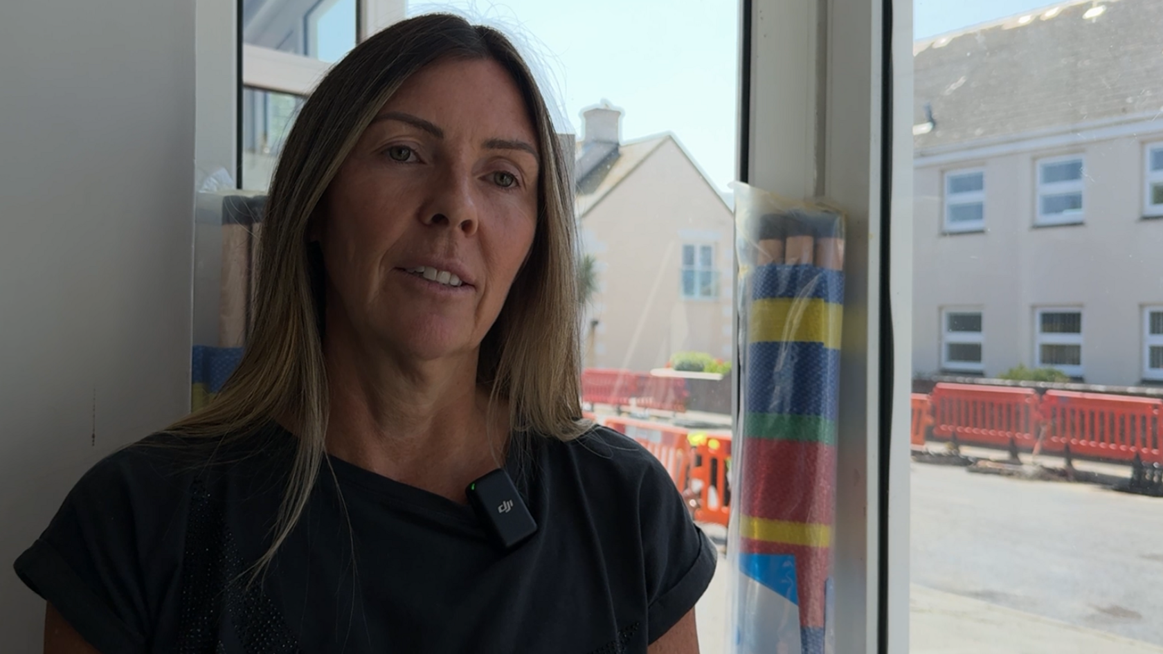 A woman standing indoors near a large window. She is wearing a dark-coloured T-shirt and has long hair. Through the window, buildings and construction barriers are visible on the street outside.