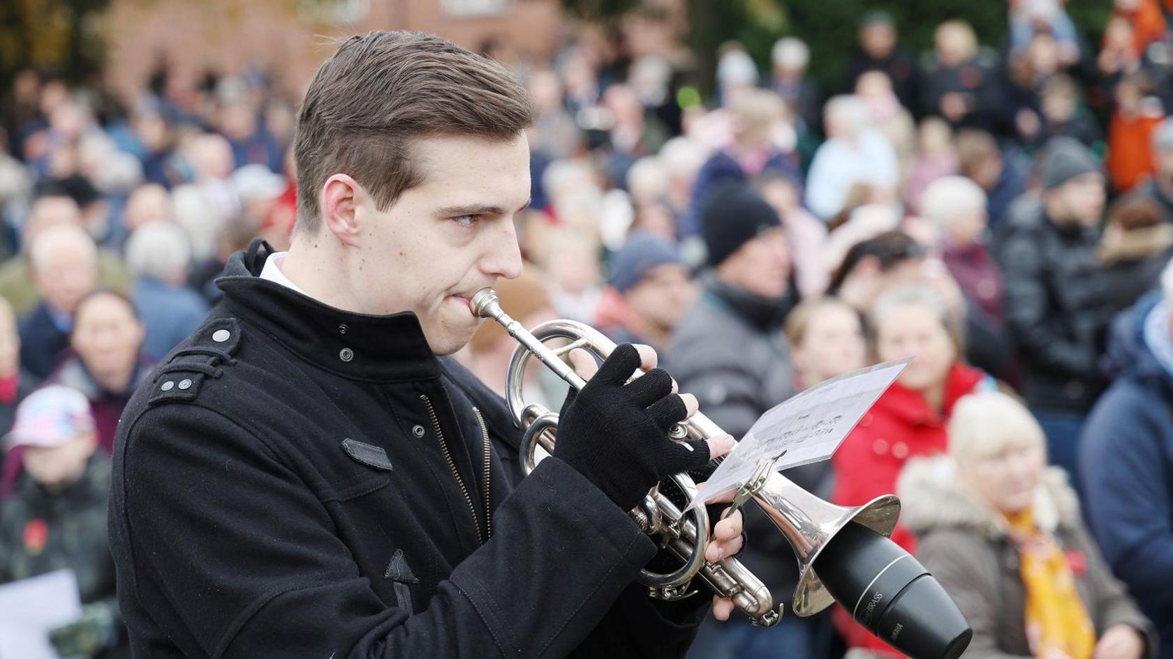 A bugler wearing a black coat and fingerless gloves plays the bugle in front of a crowd at a remembrance service