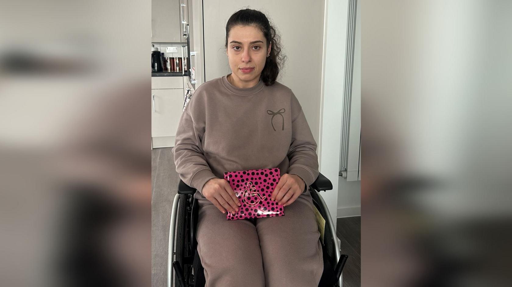 Emily Martins in a taupe jumper and tracksuit bottoms. She sits in a wheelchair and holds a pink blue badge wallet with black spots. She has dark hair in a ponytail.