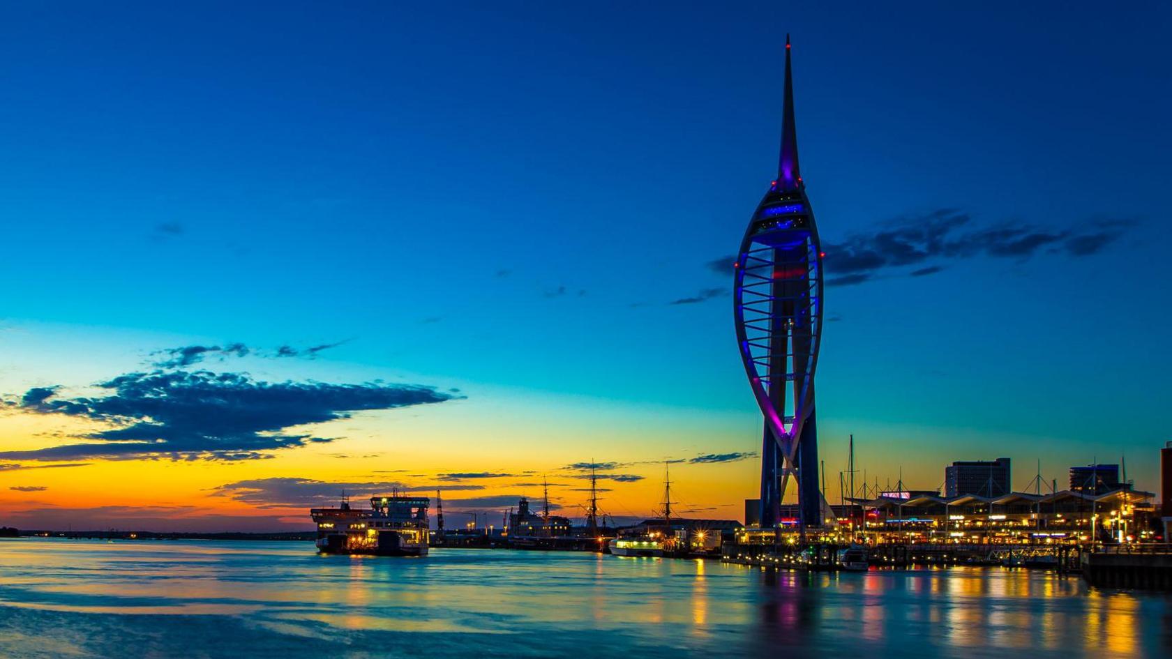 Dusk view of Portsmouth’s waterfront with the Spinnaker Tower glowing purple against a sunset sky, reflected in calm waters alongside illuminated buildings.