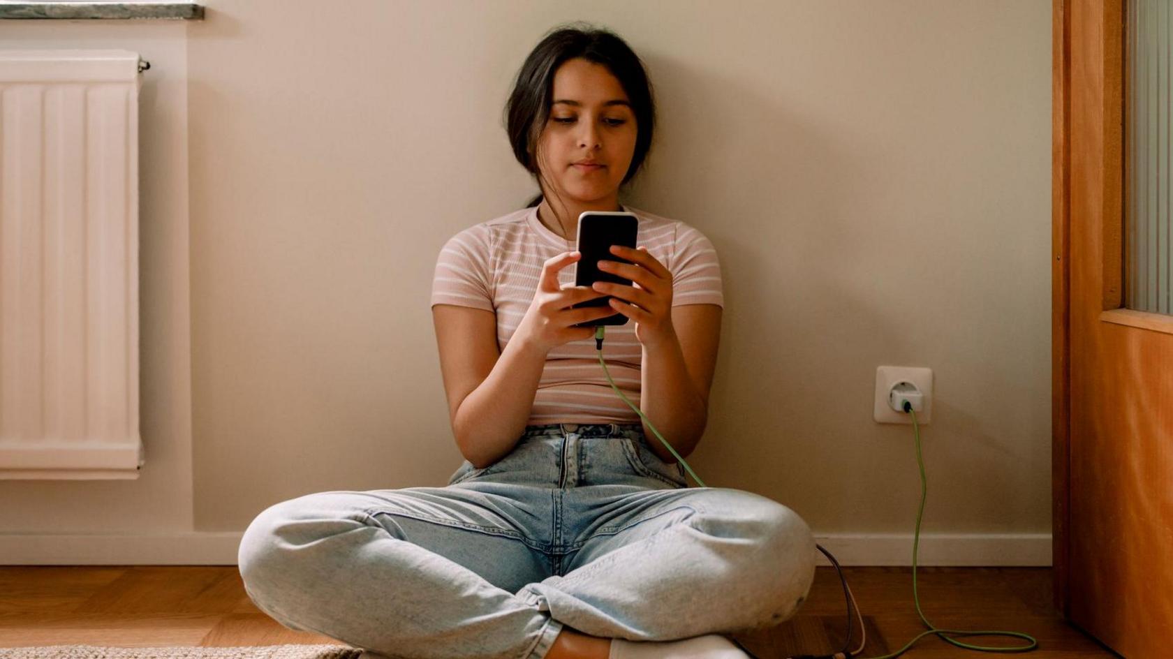 A girl wearing a pink striped top and jeans sits on the floor, looking at her phone which is plugged into the wall behind her