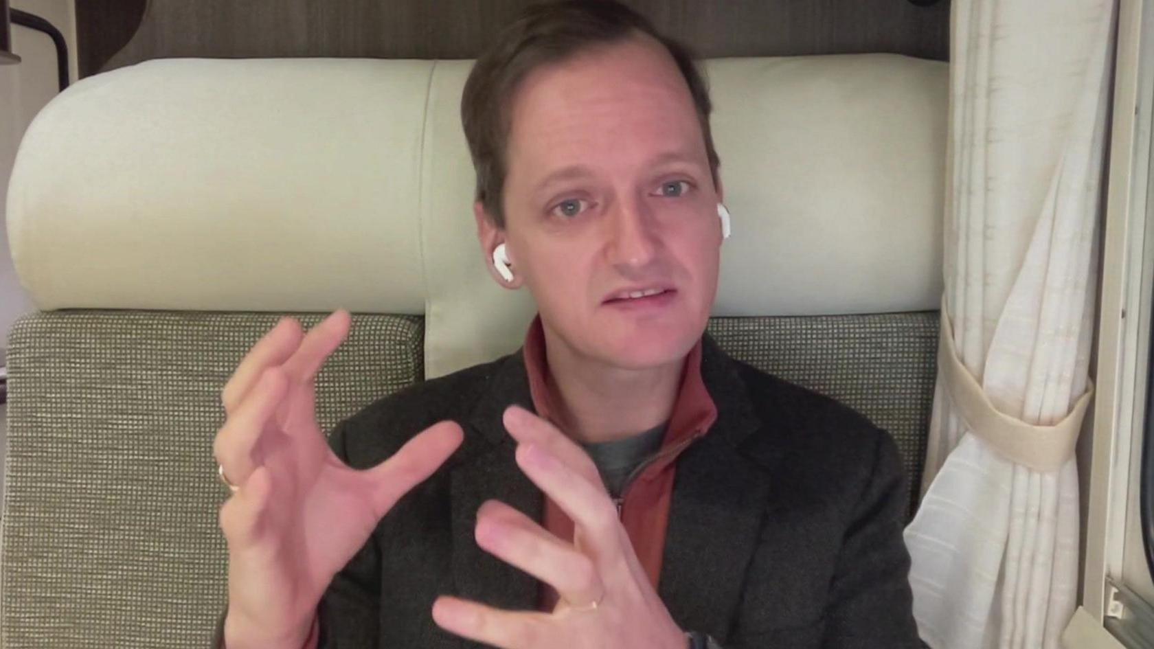 Dr Rob Salguero-Gómez sitting on a beige high-baked seat, wearing a black jacket and red shirt. He is gesturing using his hands