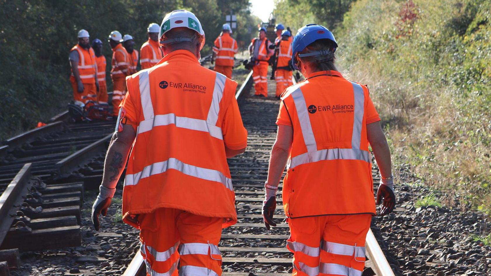 A large number of construction workers on an East West Railway line, they are all wearing orange high viz jackets and safety helmets. They are on a railway track, with gravel surrounding by trees and bushes. A man and a woman are walking away from the camera, with a group of more workers behind.