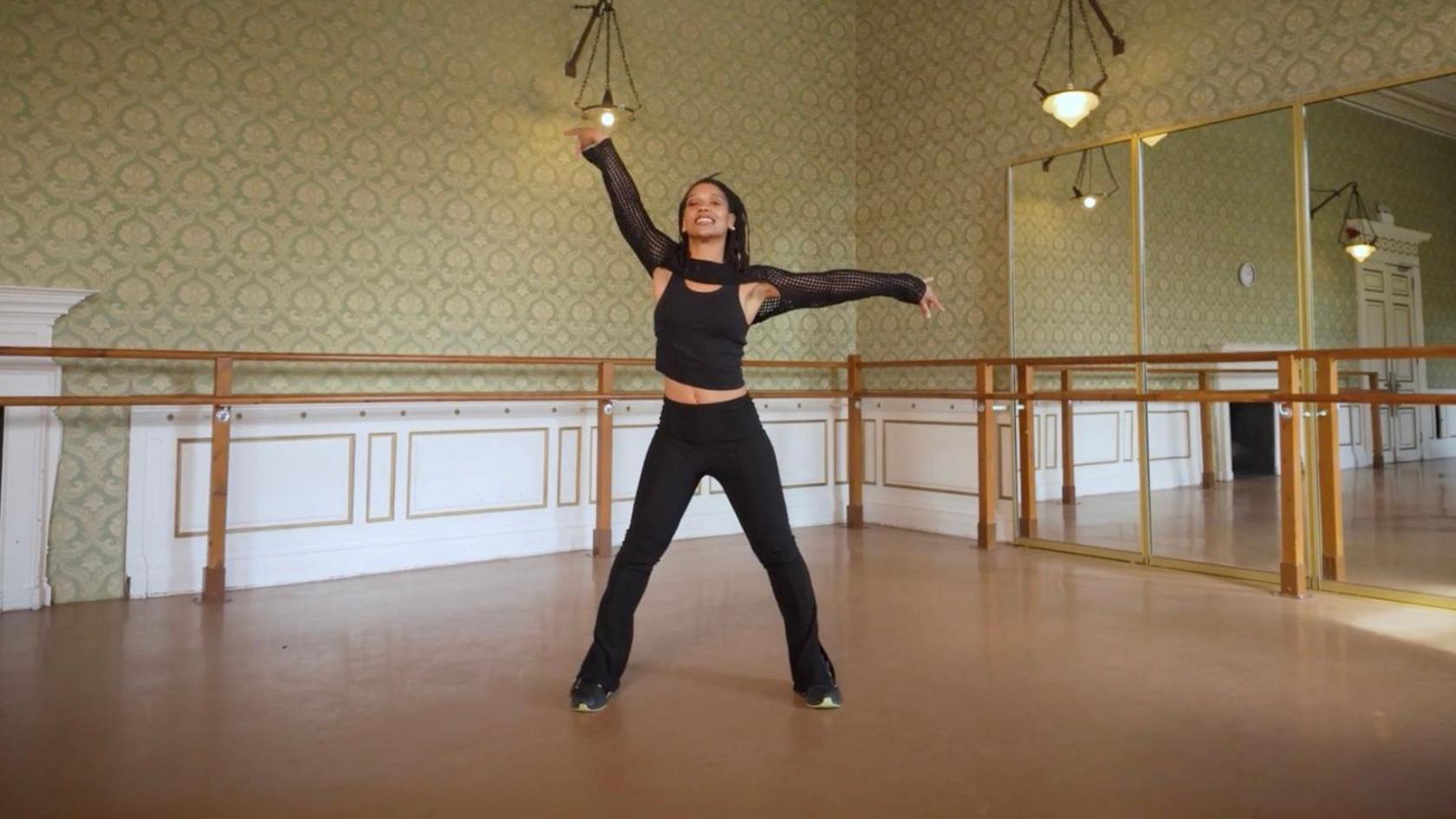 Anais Houillet performing a dance move in a room, with mirrors. She is wearing an all black dance outfit.