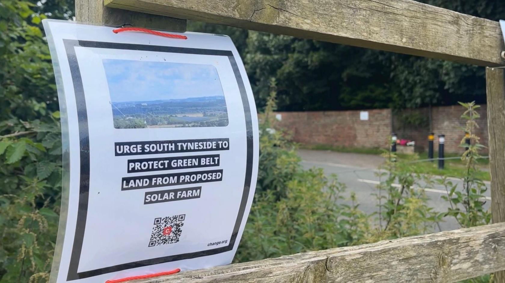 A laminated sign attached to a wooden fence. The sign reads: 'Urge South Tyneside to protect green belt land from proposed solar farm.' It also features a photo of the greenbelt and a QR code