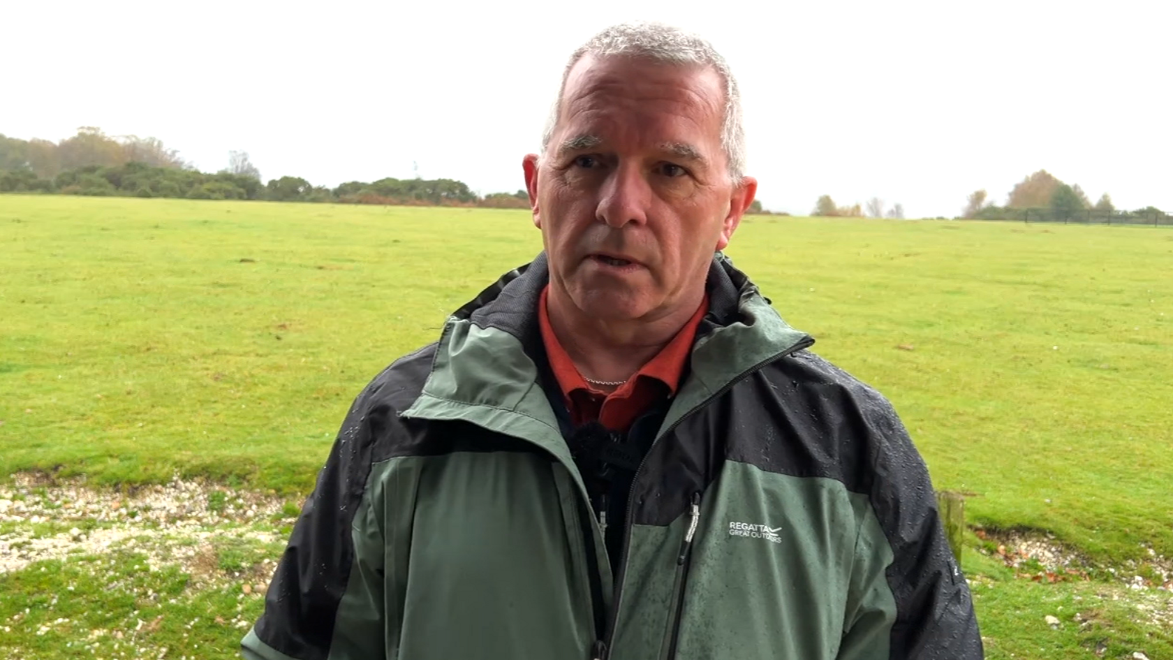 A head and shoulders photo of a man with grey hair wearing a green and black waterproof jacket. There's a large expanse of grassland behind him, with a line of trees on the horizon. The sky is cloudy and his coat is wet with rain.