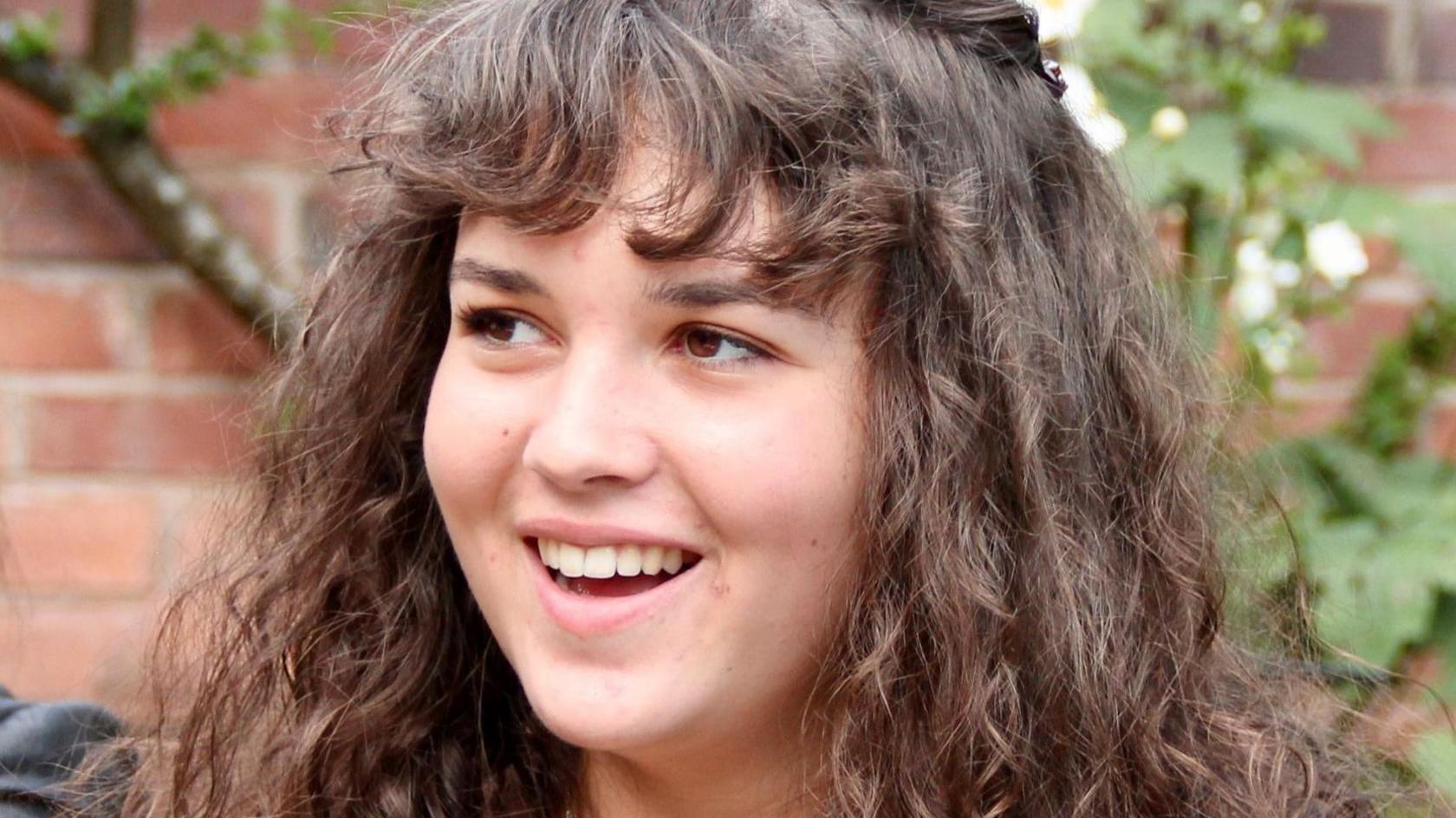 Maeve Boothby-O'Neill looking off to the left, she has her mouth open showing her teeth in a smile, she has brown curly hair and a curly fringe.