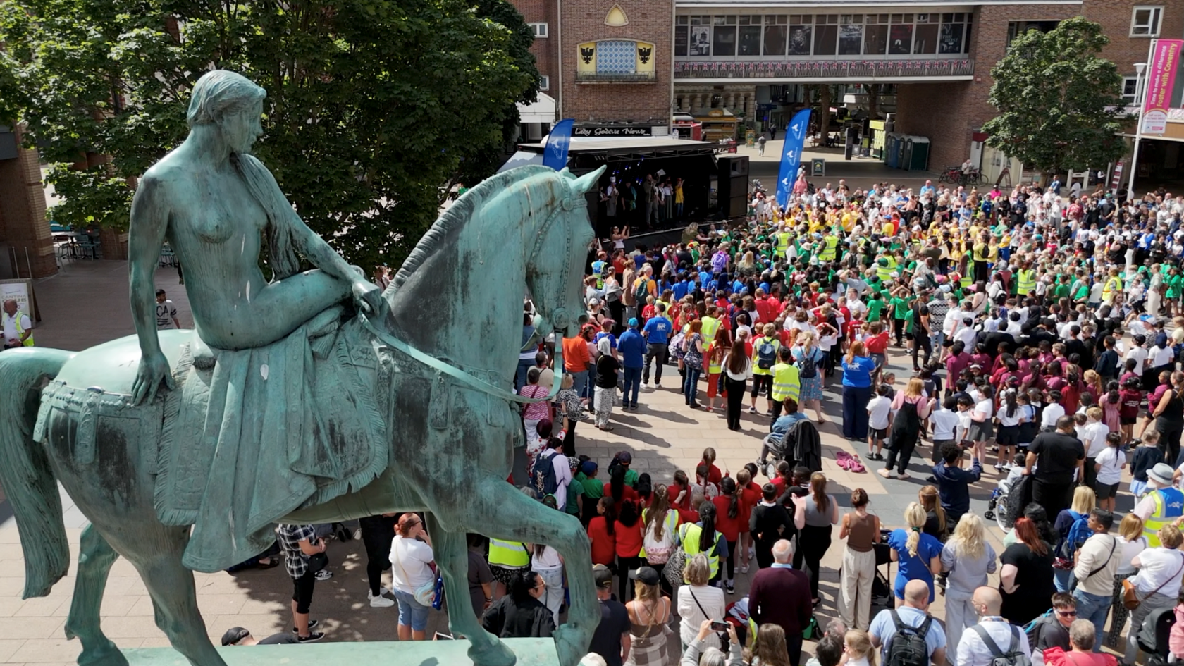 A shot from above of hundreds of people, mainly school children gathered in Broadgate main square in Coventry. A large statue of Lady Godiva on a horse is in the left of the picture and the crowd can be seen below. There is a stage in the top of the image where children are performing a musical about Lady Godiva.