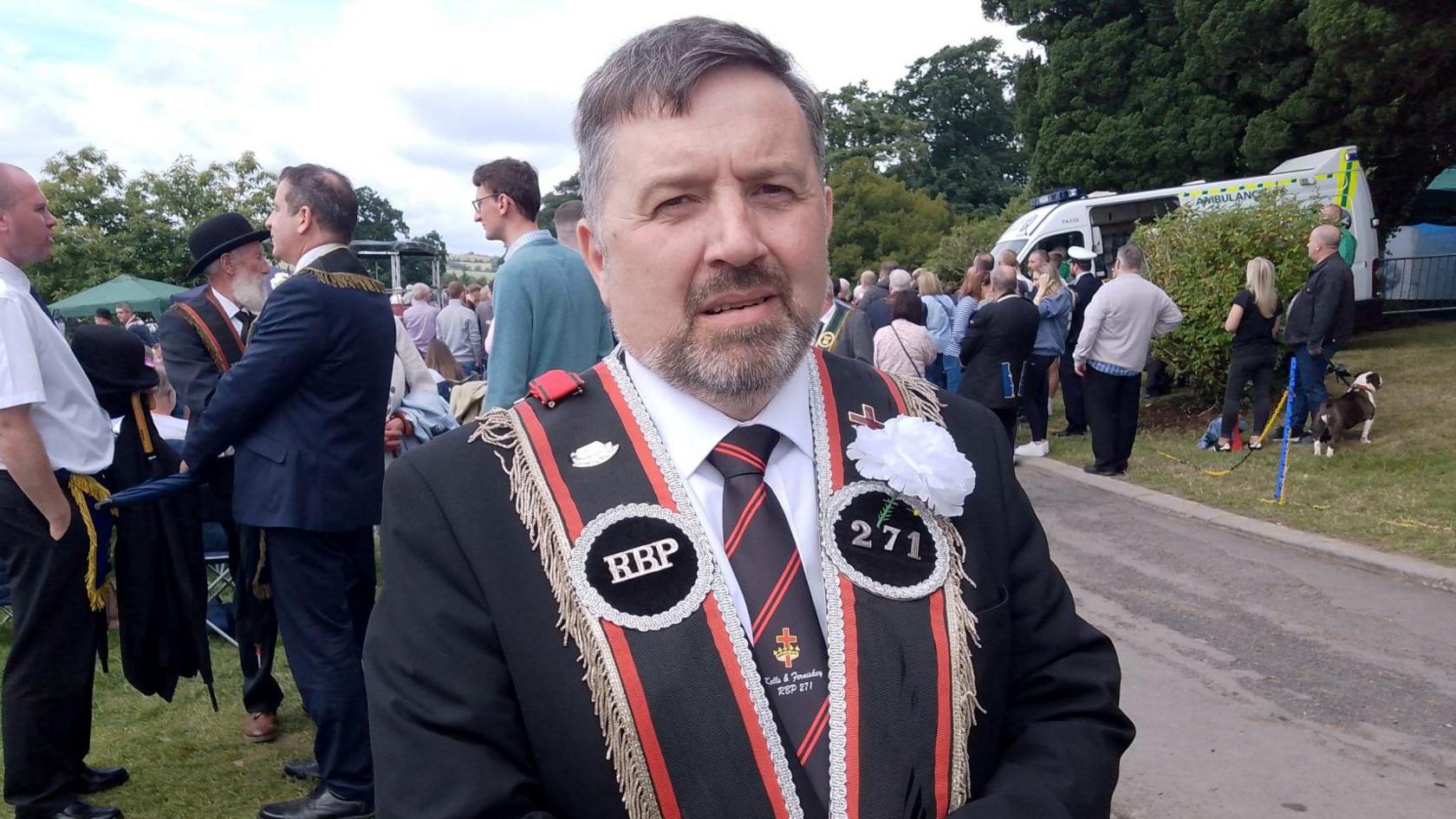 Robin Swann wears a black and red sash over his shoulders with his adorned with a white flower