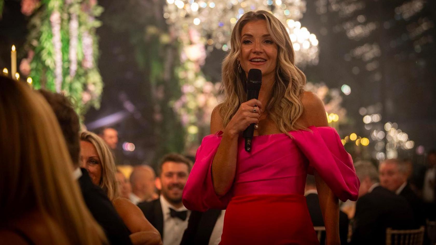 TV presenter Helen Skelton, wearing a red dress and addressing an audience with a microphone  