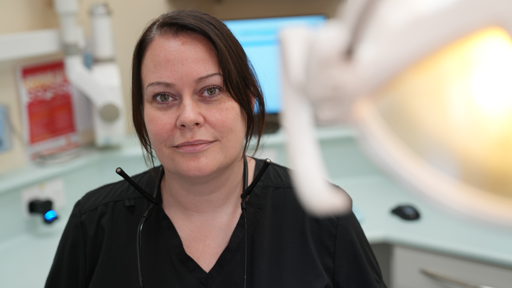 Hannah Woolnough sits in a dentist appointment room and smiles at the camera. She wears a black dentist uniform and she has dark hair that has been tied back behind her head. 