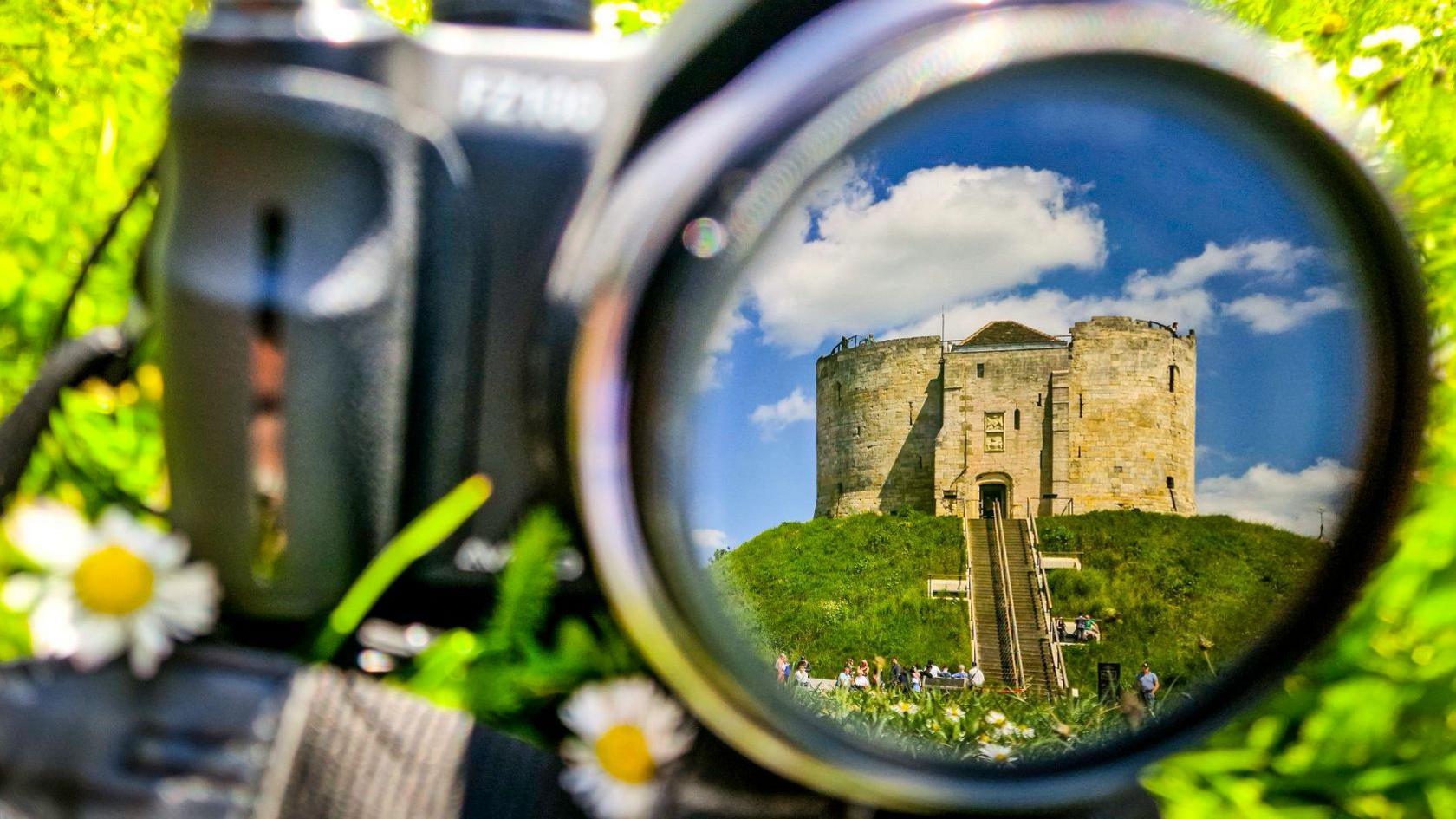 Clifford's Tower seen in the reflection of a camera lens sitting on green grass with daisies