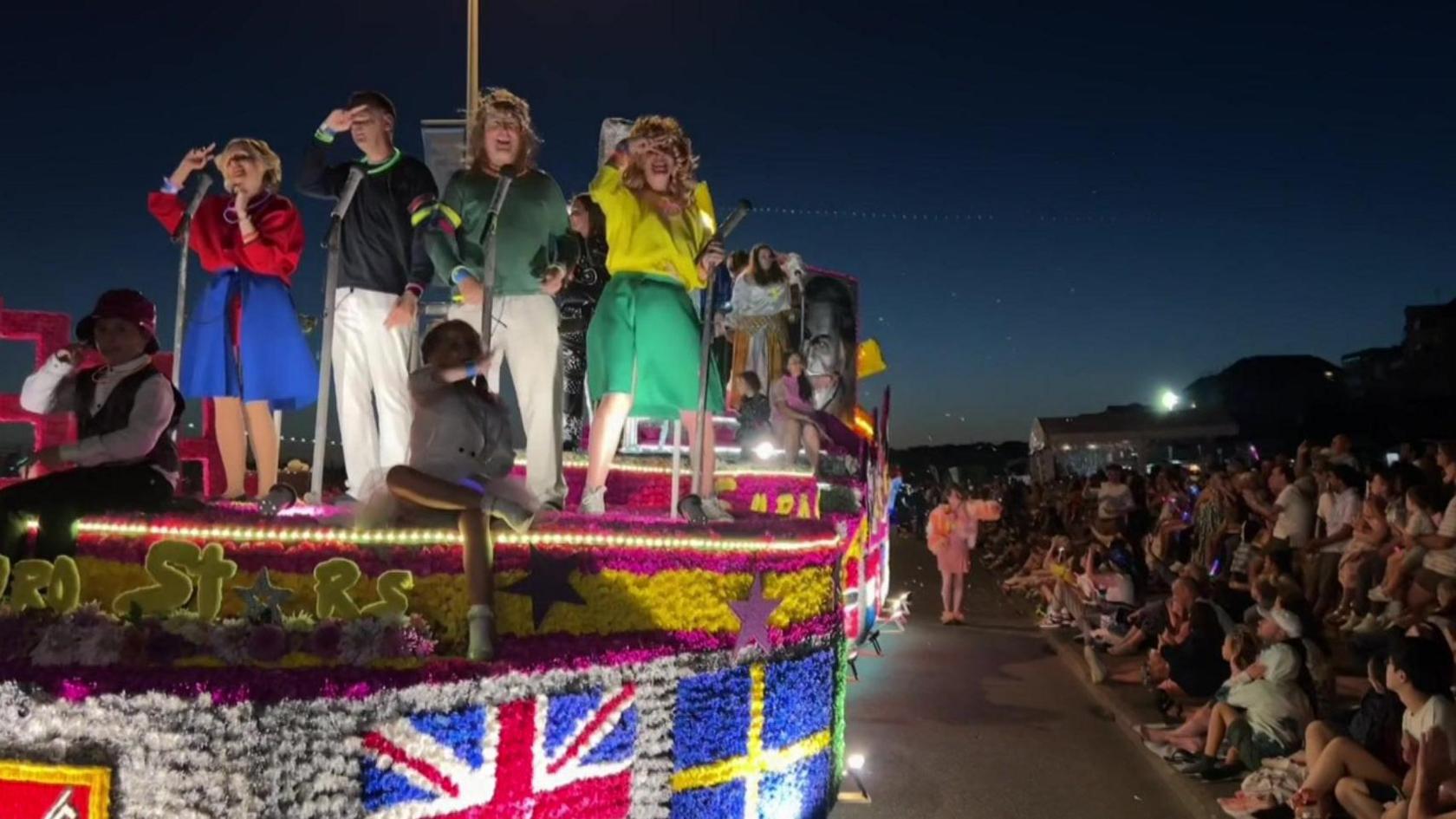 A Jersey Battle of Flowers float drives towards the camera on the left of the image while crowds line the side of the street on the right. There are several people stood on the float which is decorated in multicoloured flowers. Four of the people appear to be dressed as the 1981 Eurovision winning band Bucks Fizz and are dancing. 