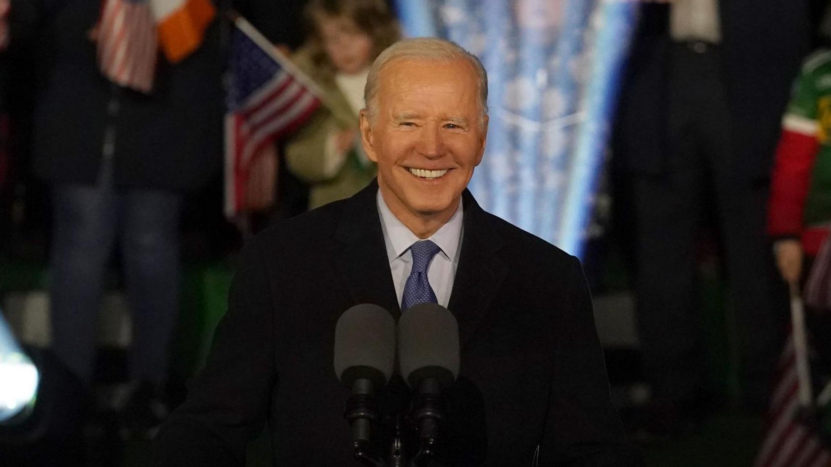 A man with white hair smiles. He is standing at a podium with an American eagle design on the front. He is wearing a suit. Behind him is a crowd, with a mixture of Irish and American flags. 