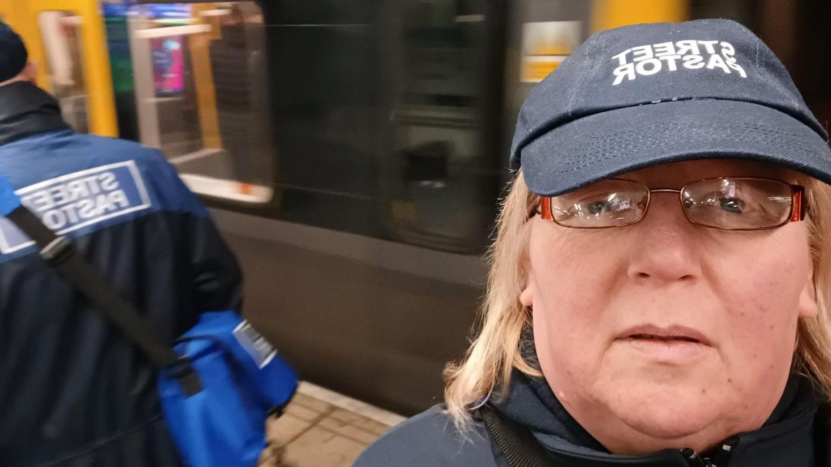 A close up picture of Julie Hudson wearing a street pastor cap. She has blonde hair and is wearing glasses. Behind her you can see the back of another volunteer.