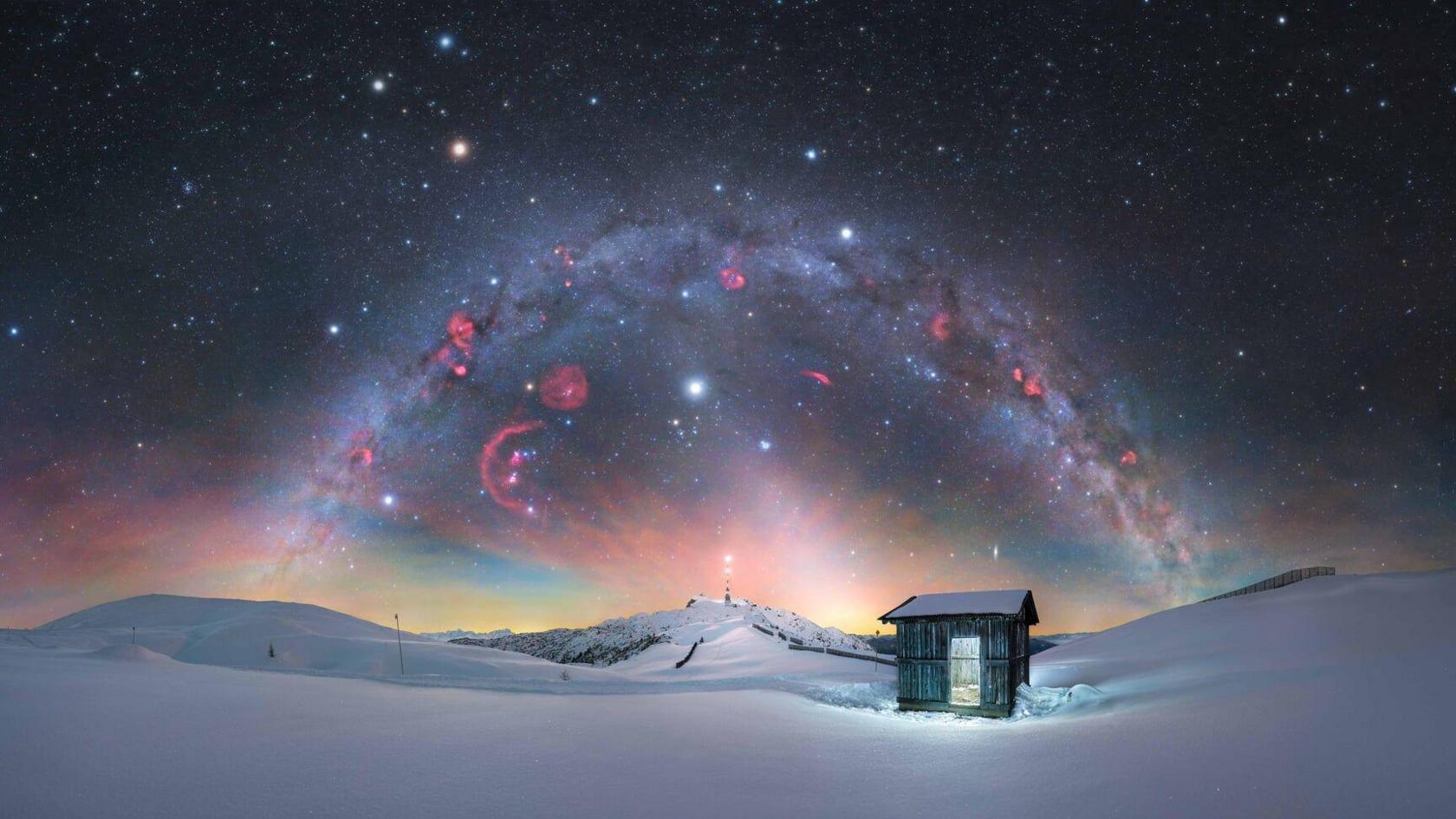 Milky Way over a snowy landscape with a cabin in the foreground.