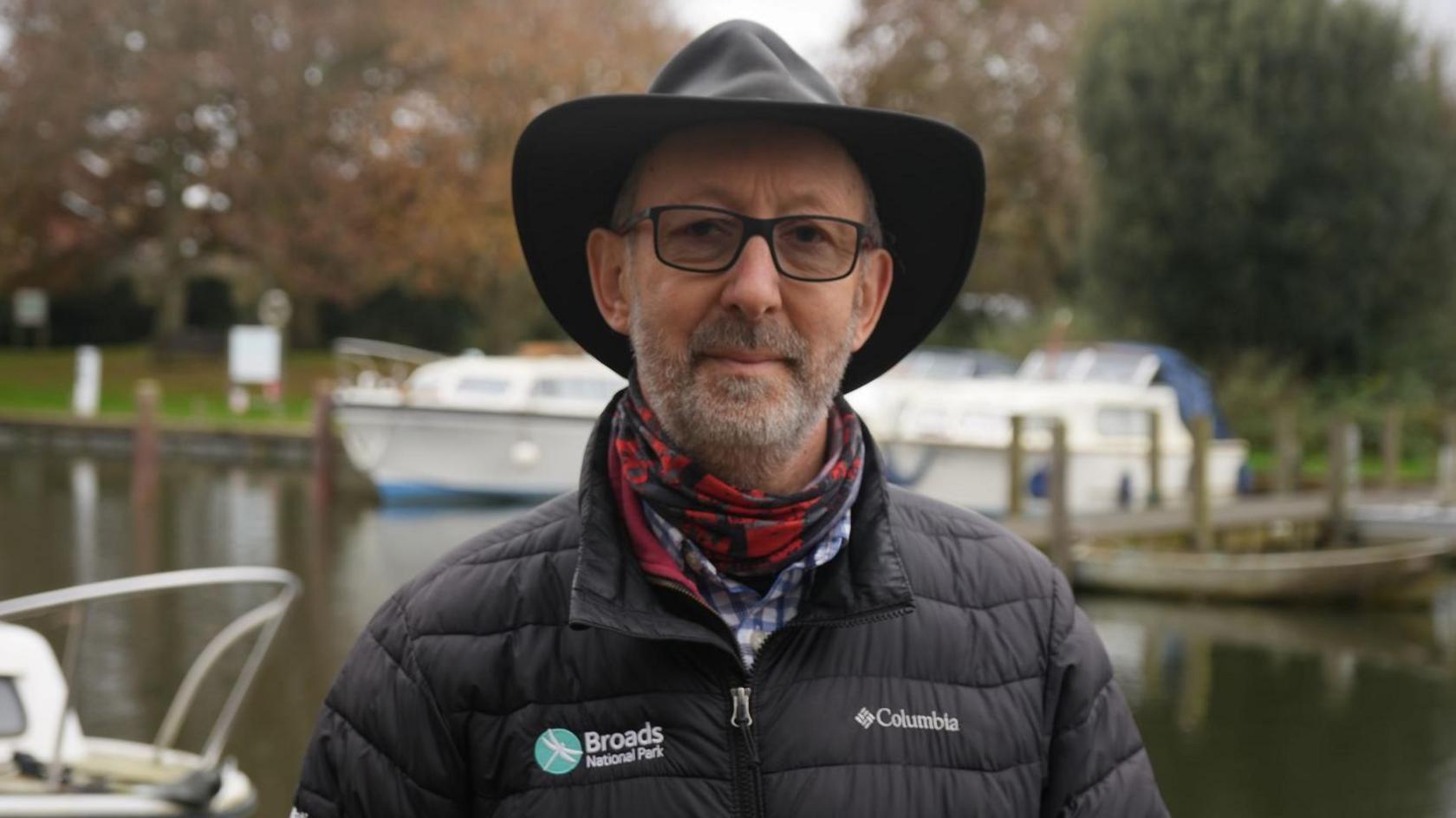 Man wears a fedora-type hat, red check scarf and black puffer-type jacket. Behind him are boats on a water course.