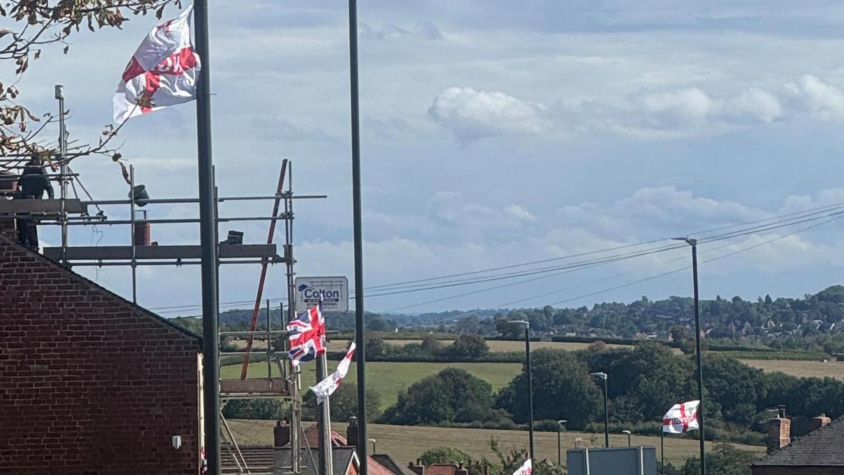 Five England flags and one Union Jack attached to lamp-posts in Codnor, Derbyshire. The road slopes down hill you can see roof tops and houses in the foreground and middle distance. Countryside visible beyond.
