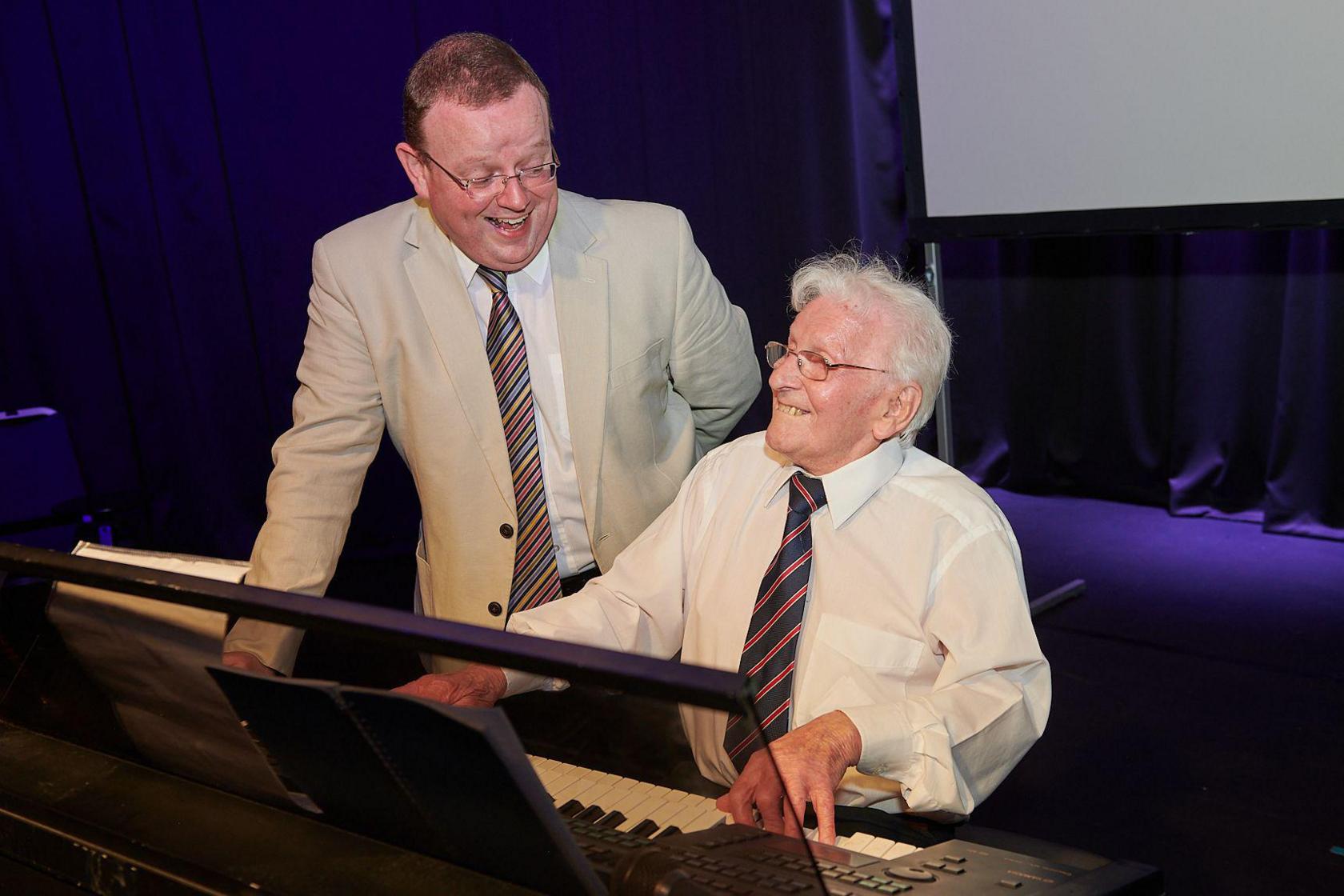 Mr Bickley sits at the organ with his hands on the keys and smiles. Mr Lloyd stands next to him and grins at him. Mr Bickley is wearing a white shirt and blue and red striped tie, while Mr Lloyd wears a cream suit jacket, white shirt and striped tie