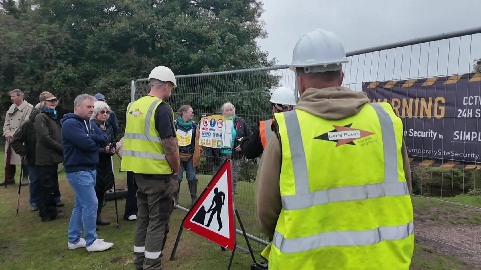 Two contractors in hi vis vests and protesters stand alongside security fencing with trees in the background