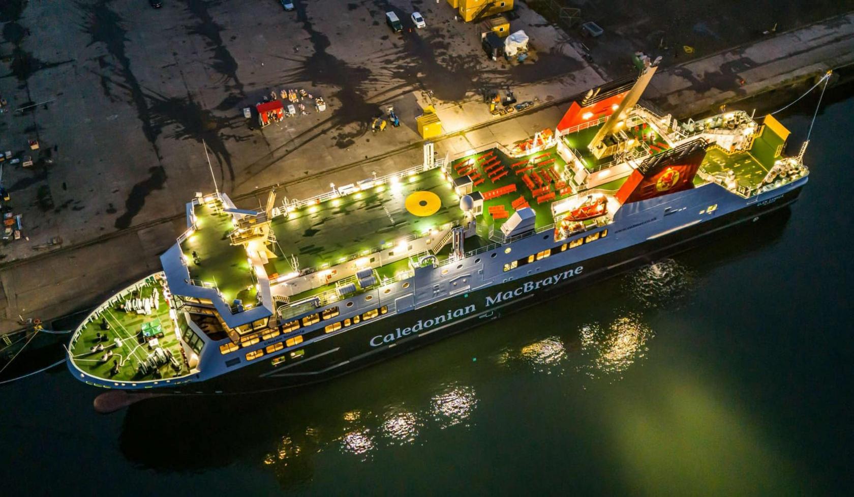 A black and white ship with green decks and red funnels, lit up at dusk beside a large concrete quay