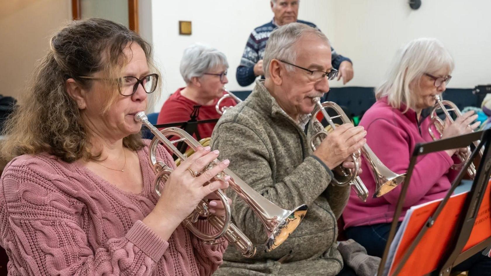 Two women and a man sit beside one another each playing a trumpet. They are all reading sheet music which is being held by a stand before them. 