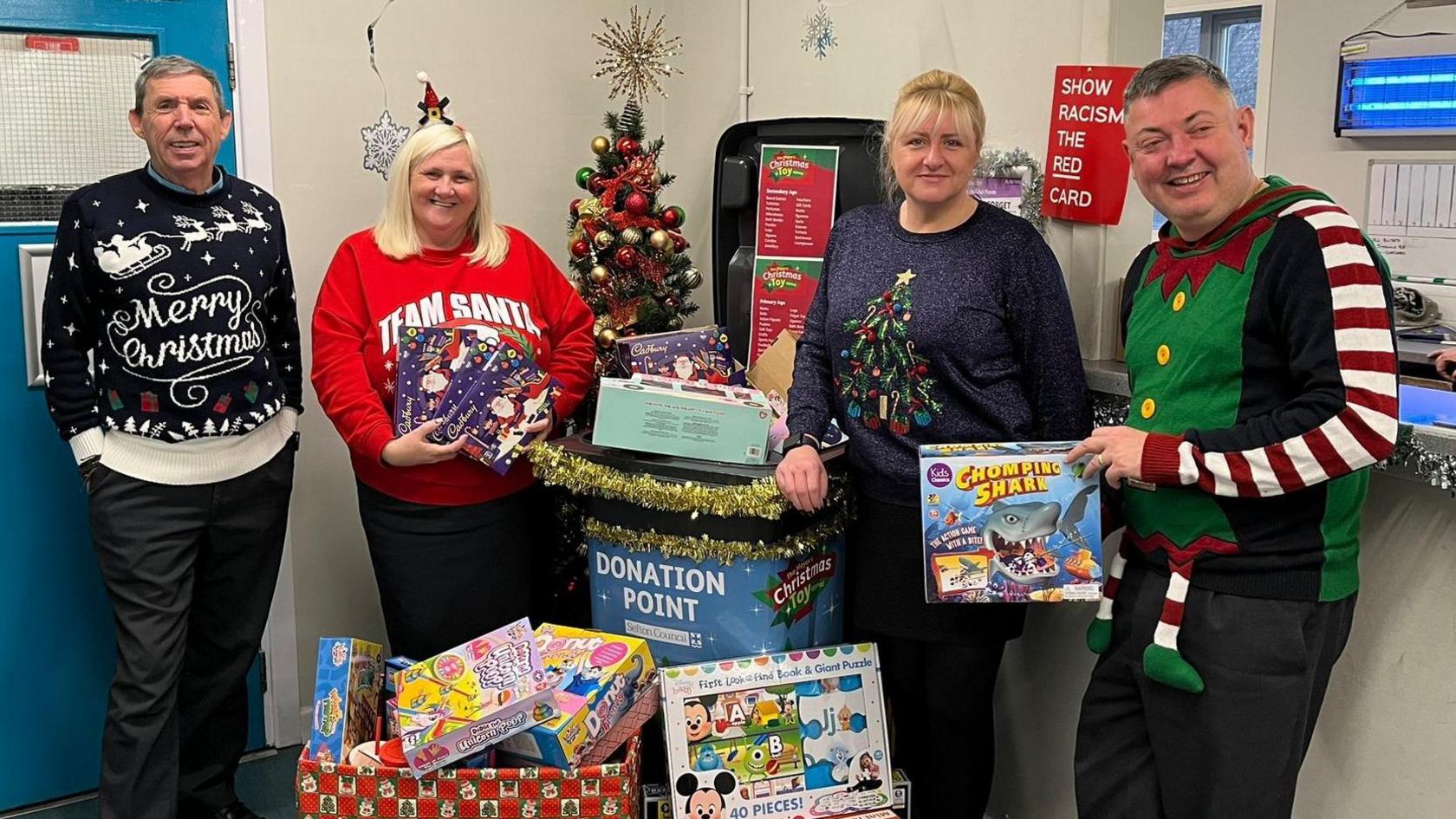 Two men and two women wearing Christmas jumpers are stood at a bus depot with toys at a donation point.