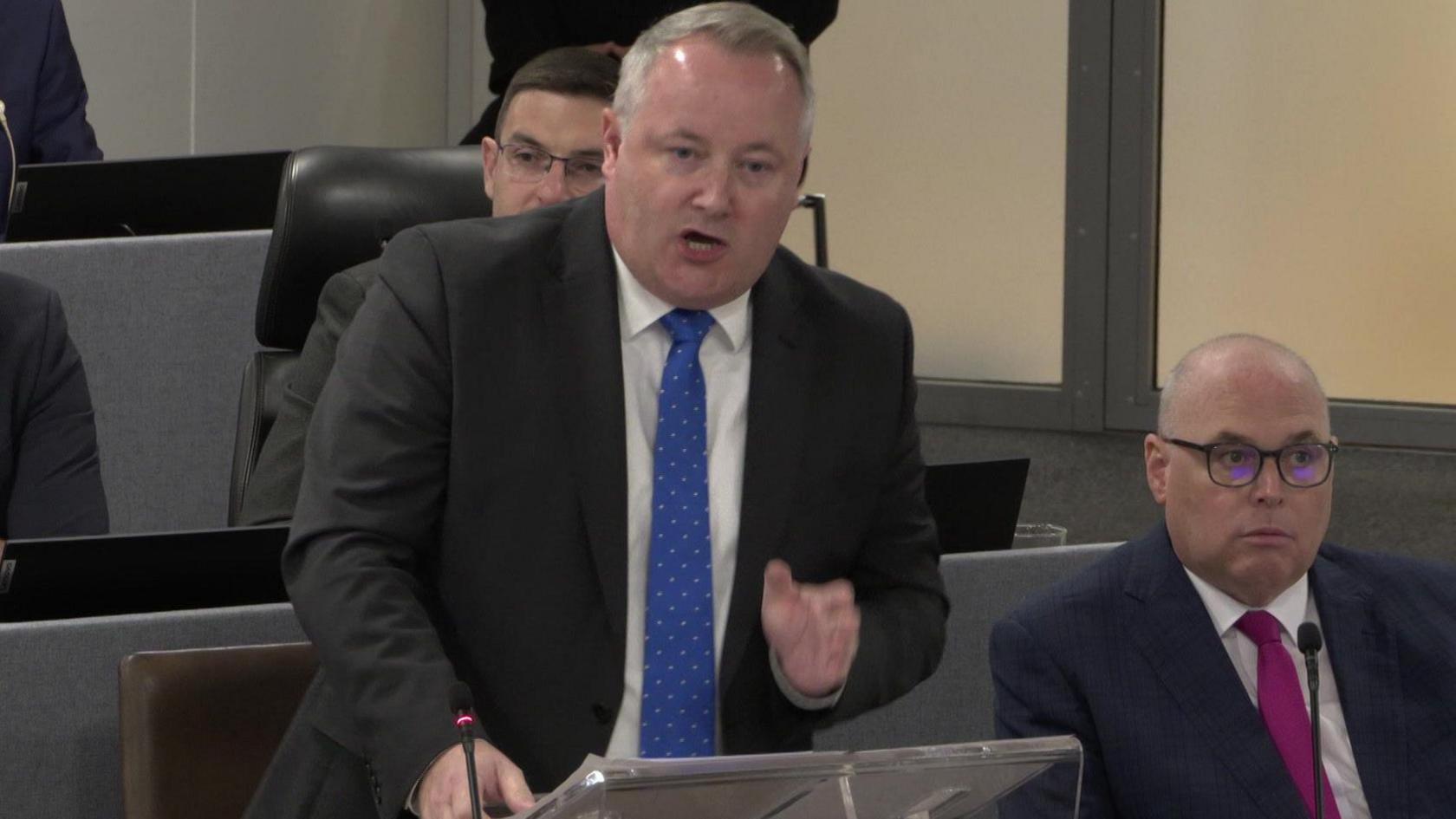 Darren Millar on his feet asking a question during First Minister's Questions in the temporary Senedd chamber in Cardiff Bay. He is wearing a blue tie with light spots, a light shirt and dark suit.