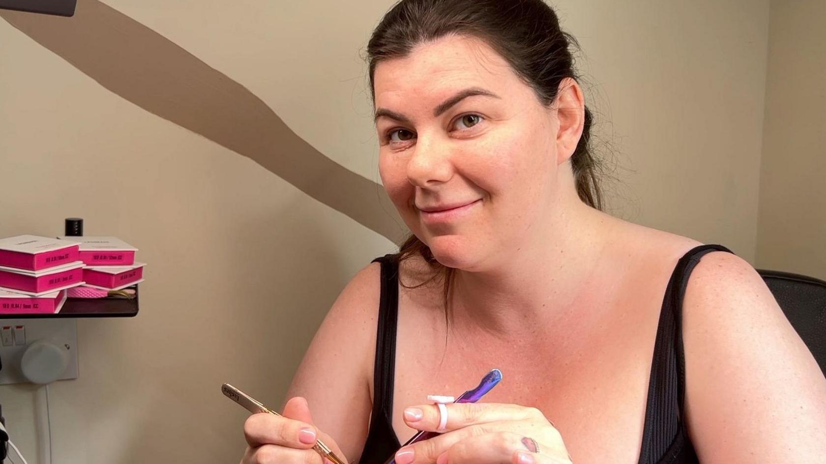 Charlotte McMahon is holding beauty salon equipment to work on a person's eyelashes. She has black hair pulled back in a ponytail and is wearing a black top