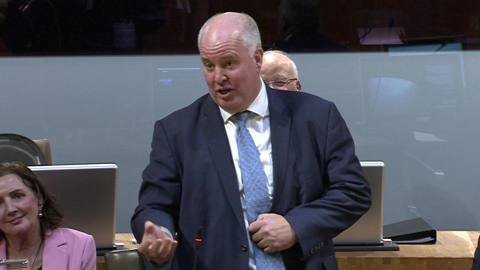 Andrew RT Davies stood in the Senedd chamber, wearing a blue suit and blue tie, surrounded by other Conservative Members of the Senedd.