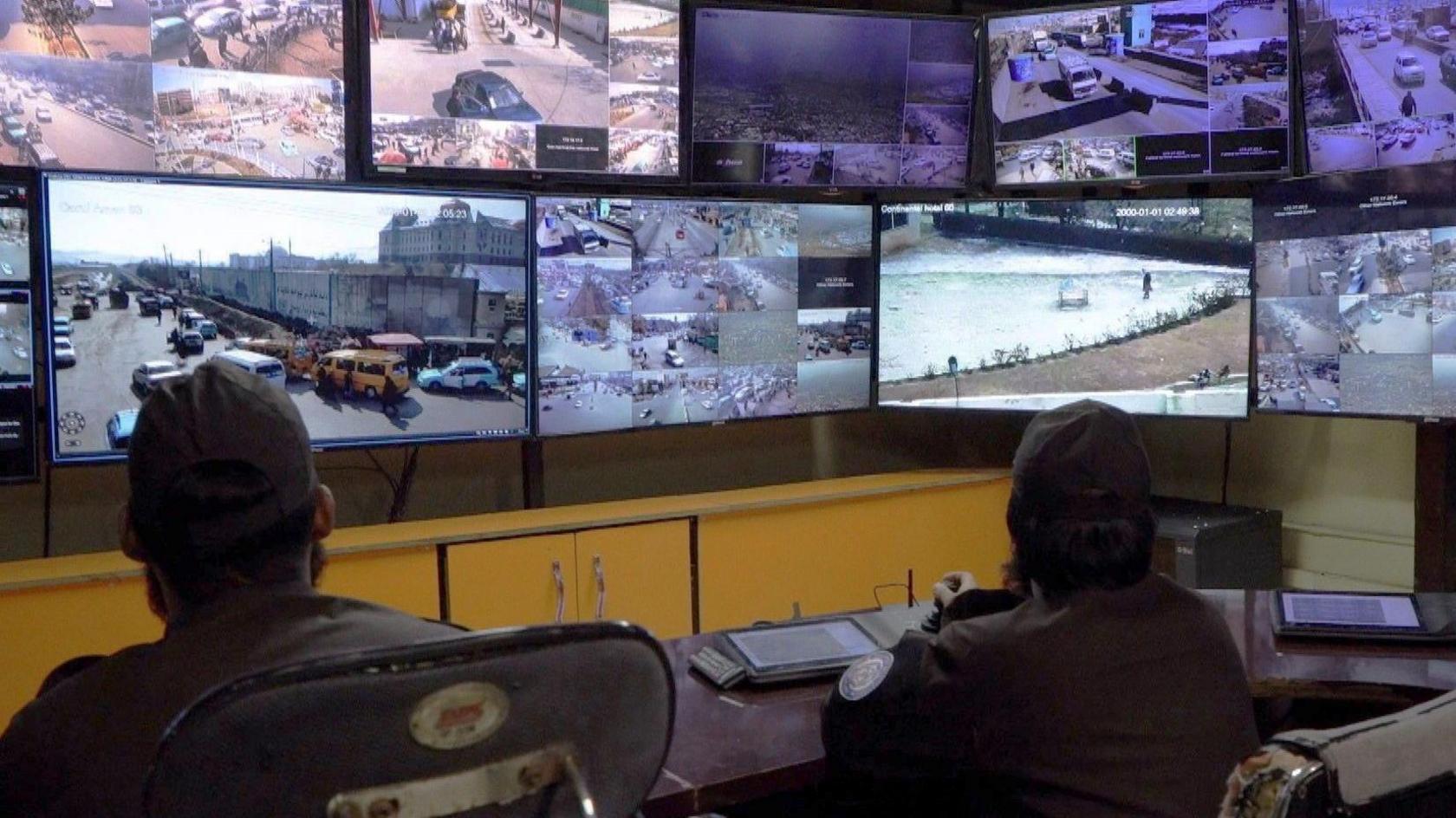 Two men are seen from behind looking at a bank of screens