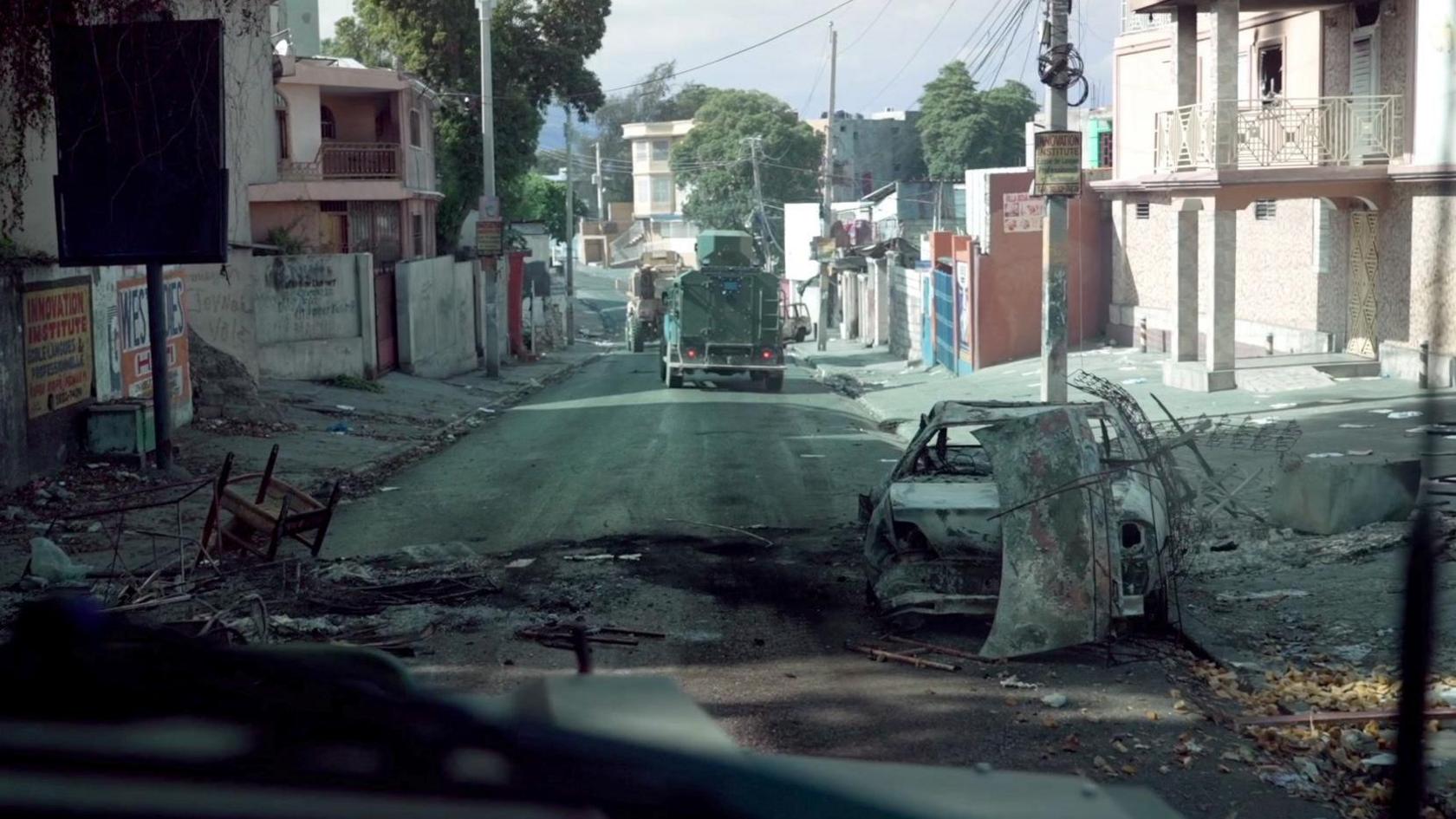 Burnt out vehicles litter a street in Port-au-Prince, an armoured vehicle can be seen driving ahead. 