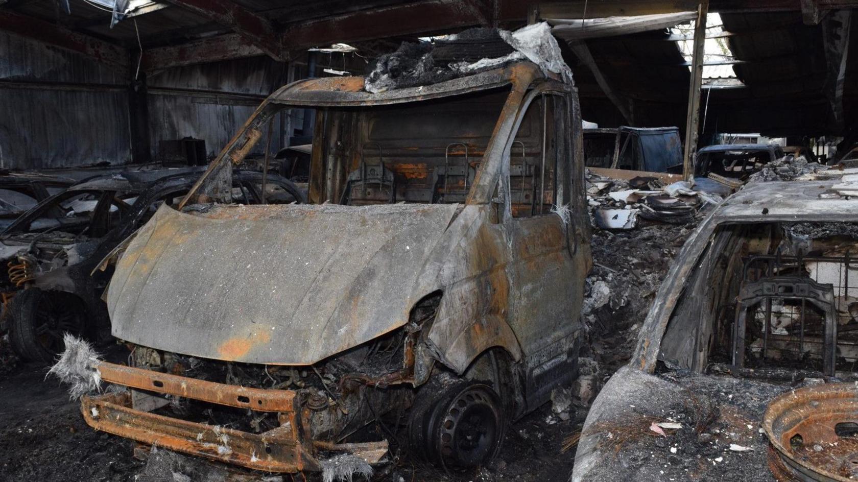 A burnt out van is sitting in a charred warehouse surrounded by other burnt vehicles