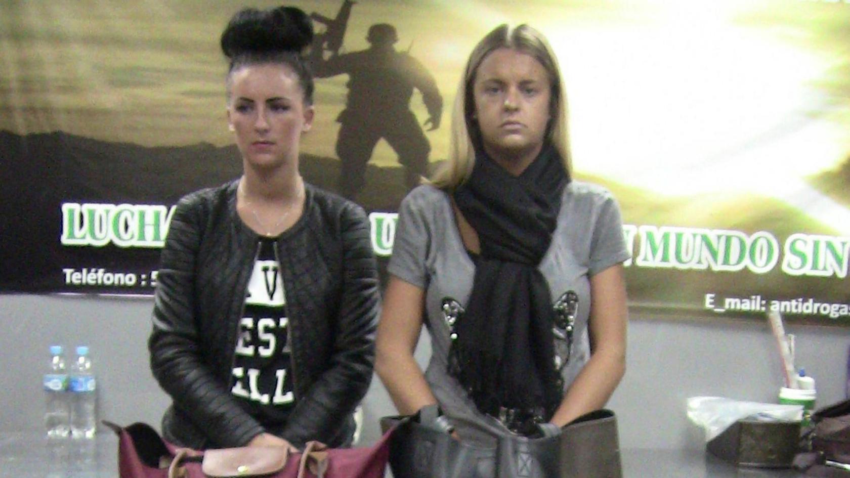 A younger Michaela McCollum, with dark hair in a large bun, stands holding her hands behind her suitcase in Lima Airport. Beside her is Melissa Reid, who has longer blonde hair and is looking at the camera with a neutral expression on her face.