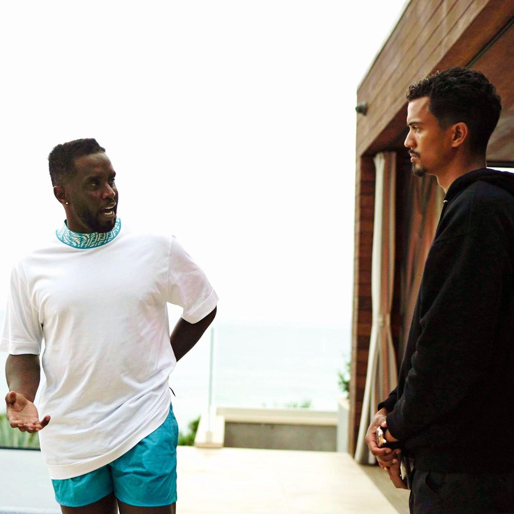 Sean Combs (left) and Phil Pines (right) talk outdoors near a building with an ocean view in the background; Combs wears a white t-shirt and blue shorts, Pines is in a black hoodie.
