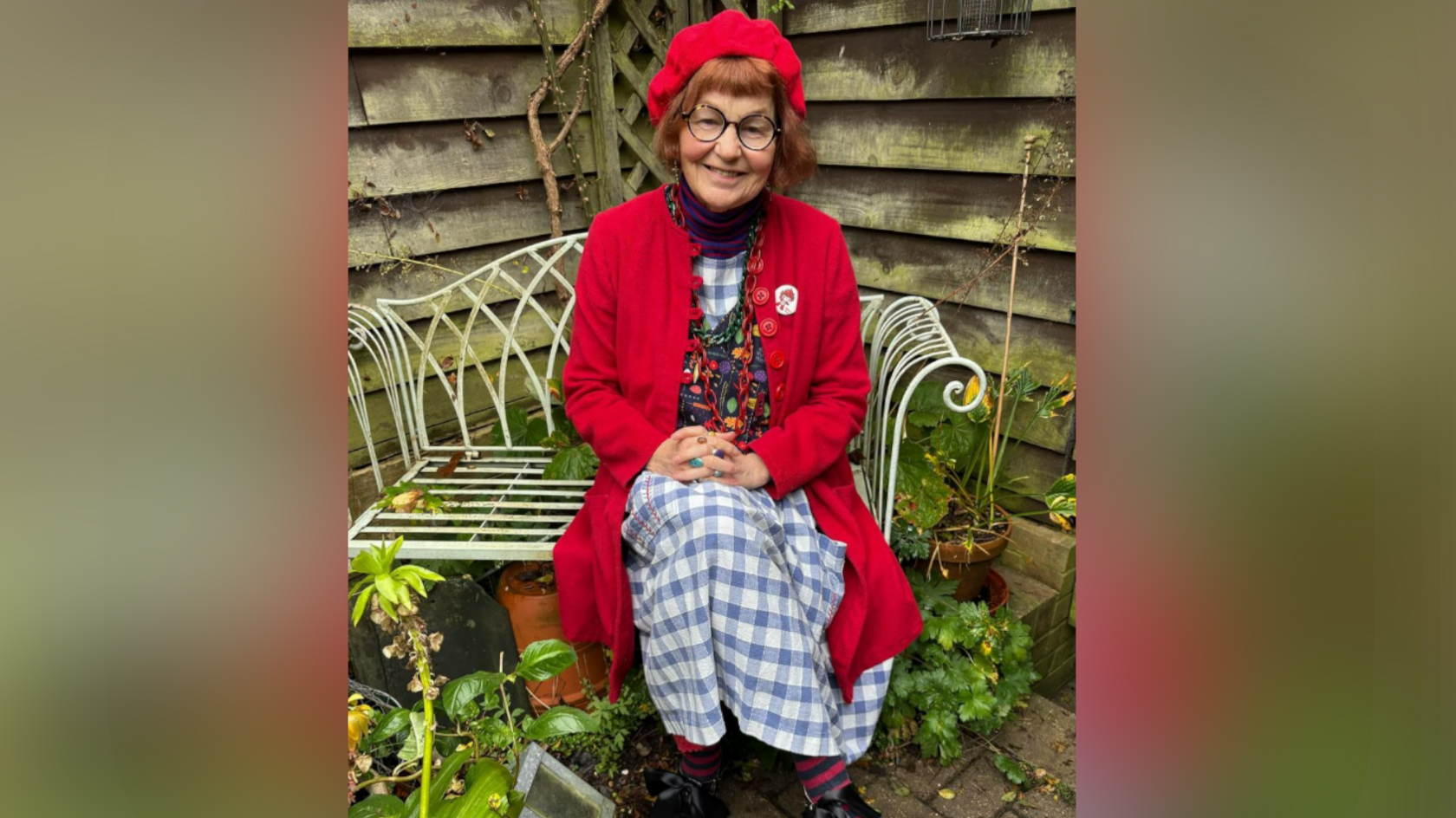 Marcia Riddington wears a red hat with a long red cardigan. She has red hair and black glasses and is sitting on a garden bench. Beneath her cardigan she is wearing a chequered blue and white dress with a multi-coloured vest over it. 