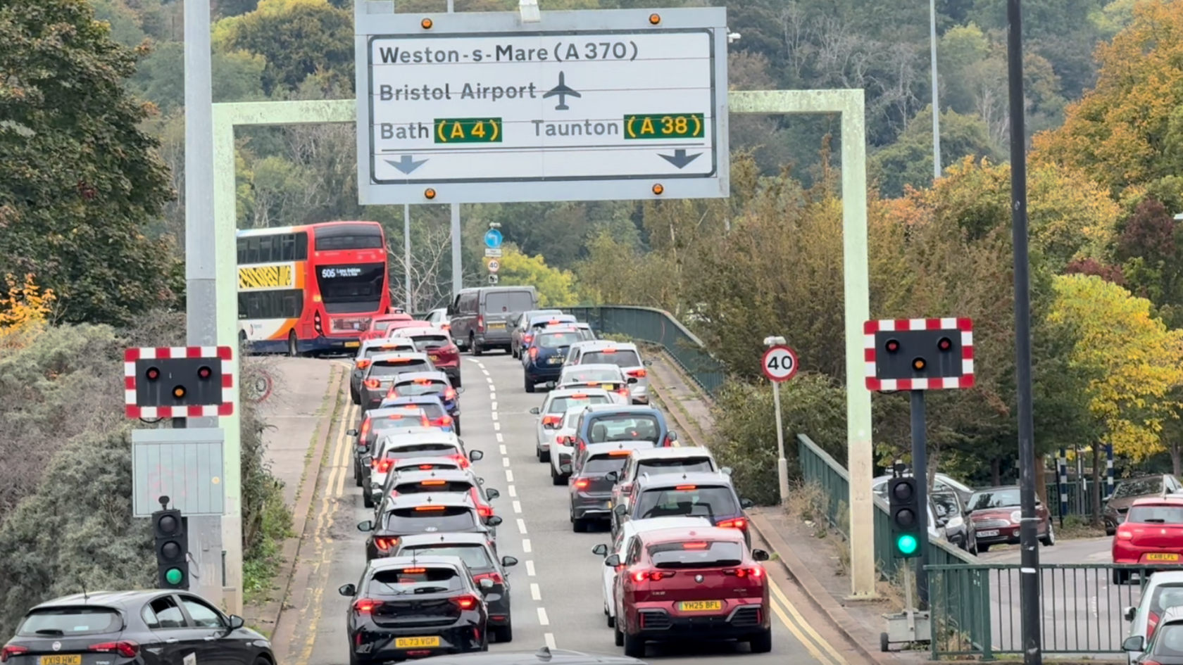 Two lanes of traffic are queuing on the slip road onto to the Plimsoll Swing Bridge/Brunel Way. A traffic sign is showing directions to Weston-super-Mare, Bristol Airport, Bath and Taunton. Trees around the road have orange and green leaves. 