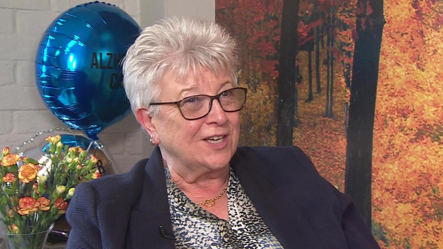 Maggie Bennett, an older lady who is wearing a blue blazer. She is sat down with a balloon and a bunch of carnations behind her