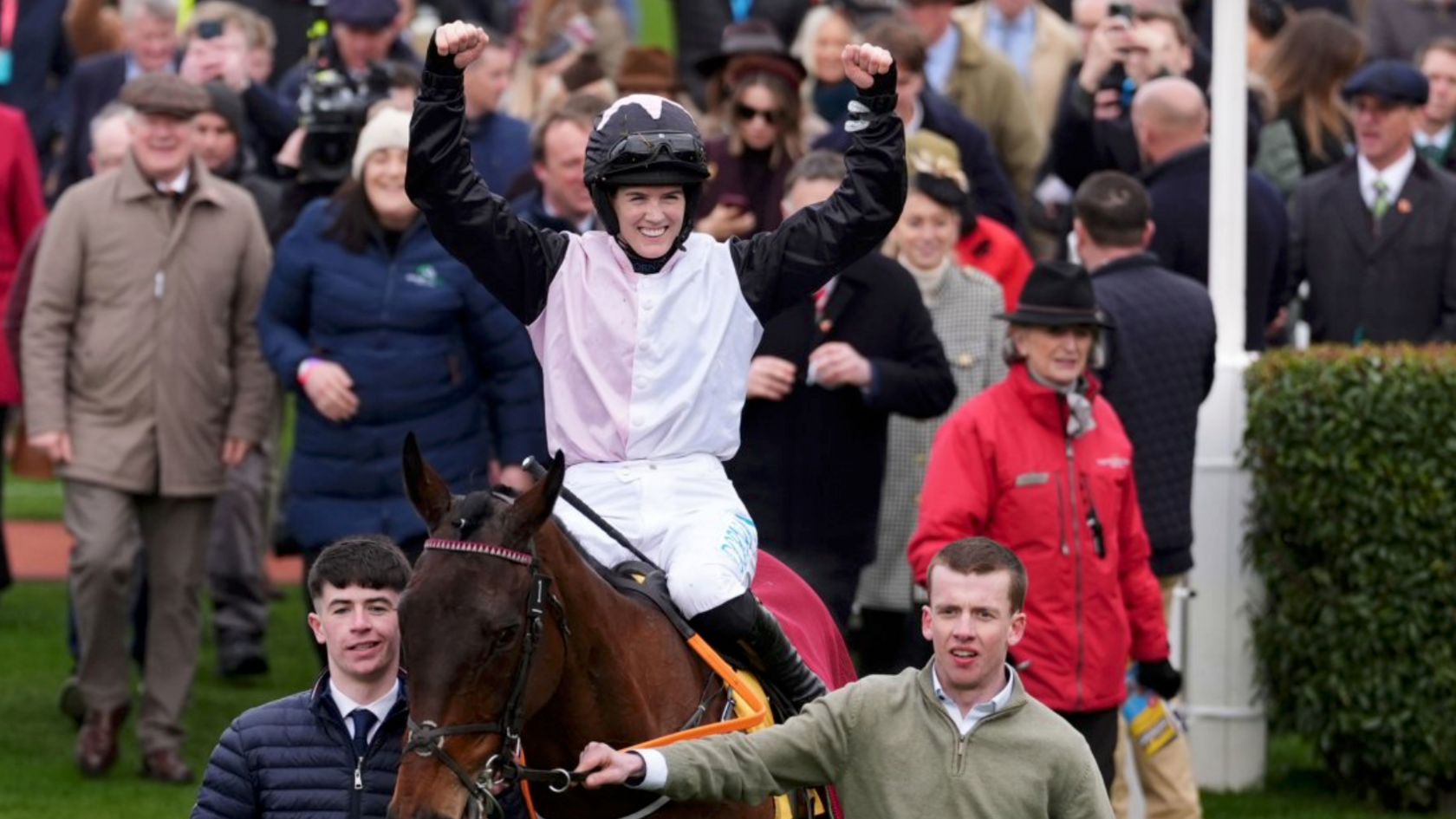 Rachel Blackmore is smiling with her arms up sitting on a brown horse. She is wearing a black white and light pink silk with a black helmet decorated with a pink star.