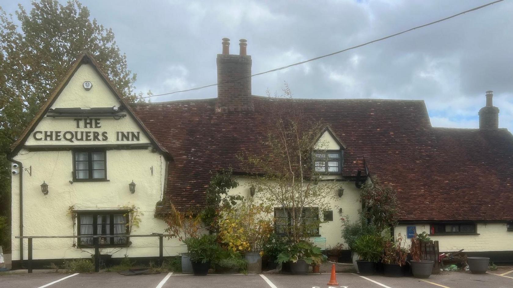 The Chequers Inn before it was burned down. It is an old building with a cream facade and large sloped roof made of red tiles. There is a chimney in the middle and pots of shrubs and trees outside. There are car parking spaces in front of the pub.