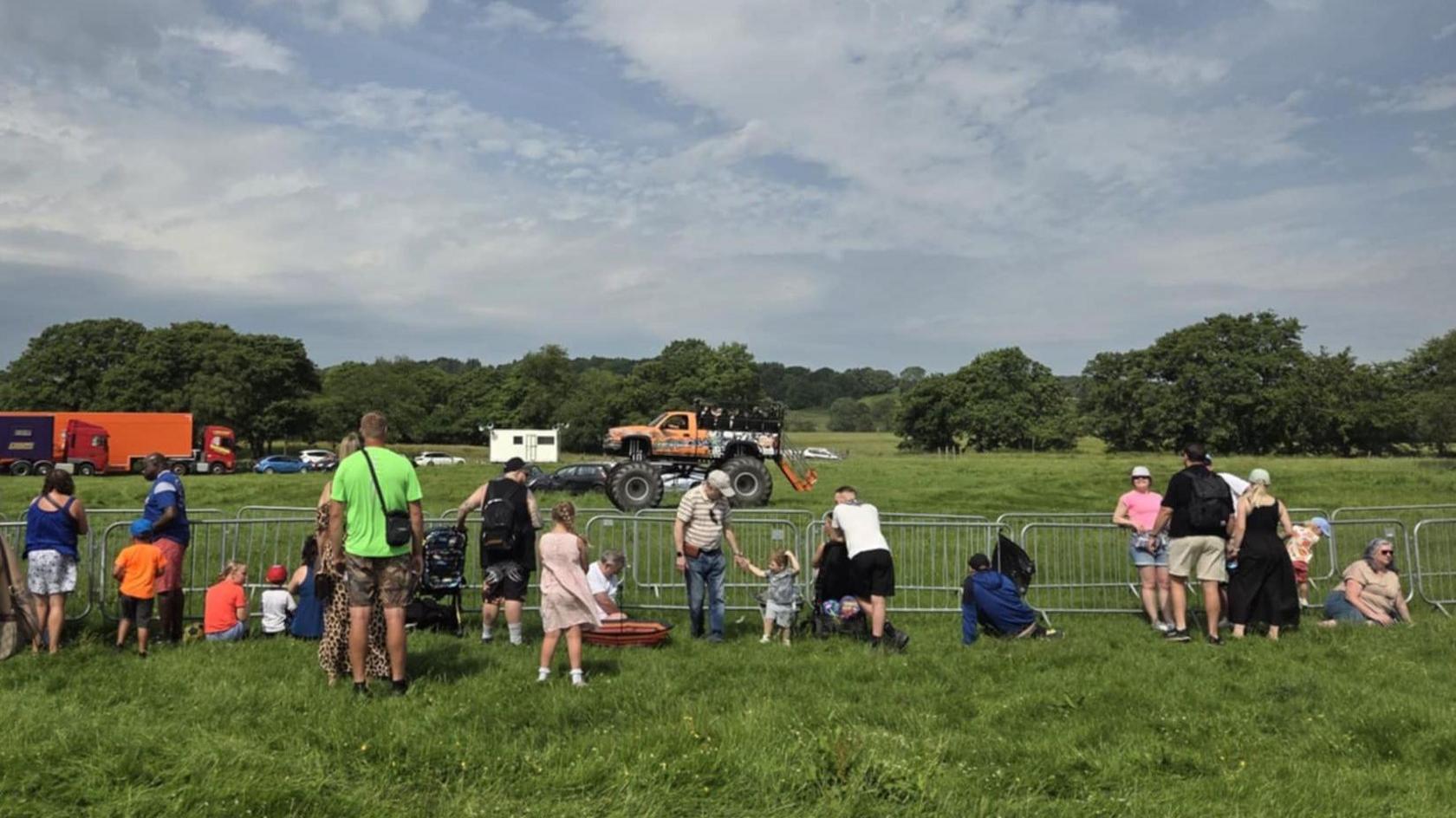 Families are standing next to an iron fence where an orange monster truck is sitting on a field.
