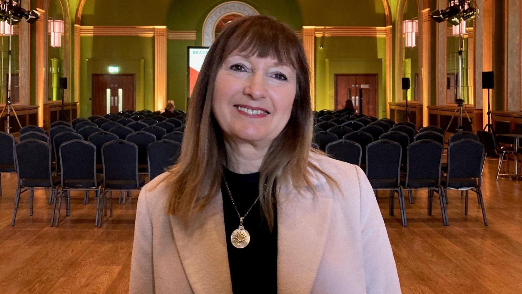 A woman with long dark hair and wearing a pink jacket and black jumper stands in a room with a high ceiling and golden archways.