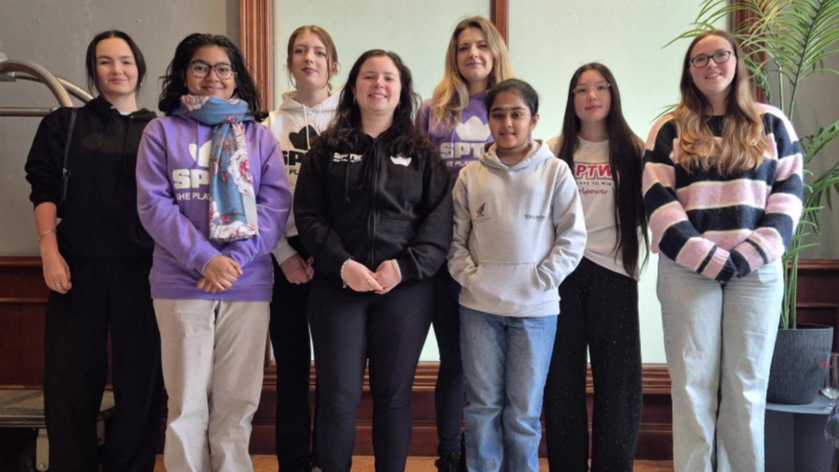 Eight teenage girls of various heights and ethnicities standing in a line