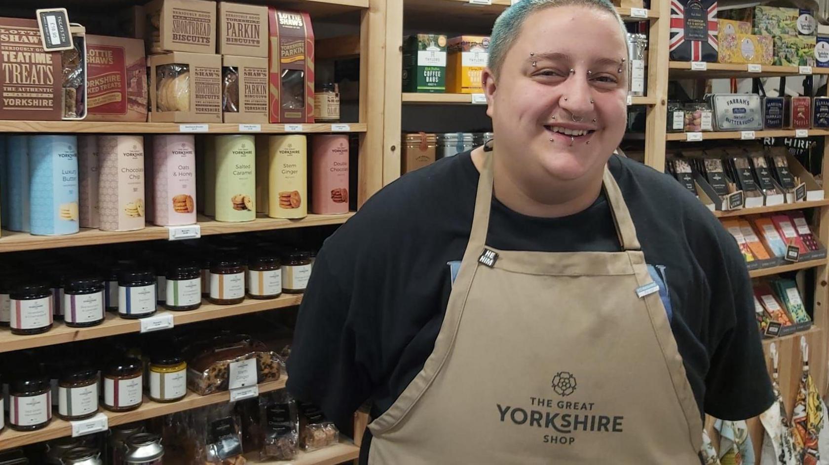 A man with multiple piercings and blue hair stands in front of shelves of chutney wearing a brown apron.