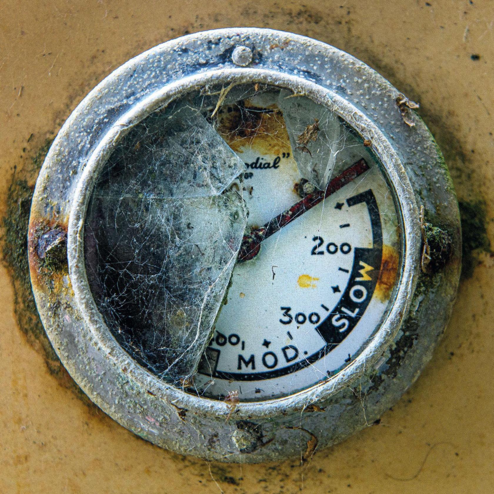 A dial on a yellow wall. The glass front is smashed and numbers and the word "slow" are visible on the dial.