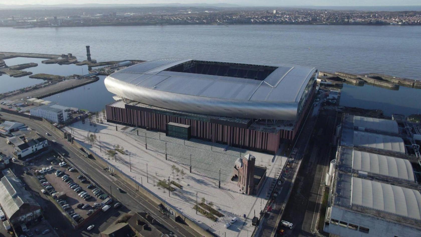 An aerial view of Everton Football Club's new Bramley-Moore Dock stadium. It is a large silver structure with a curved roof. The stadium is on the banks of the River Mersey.