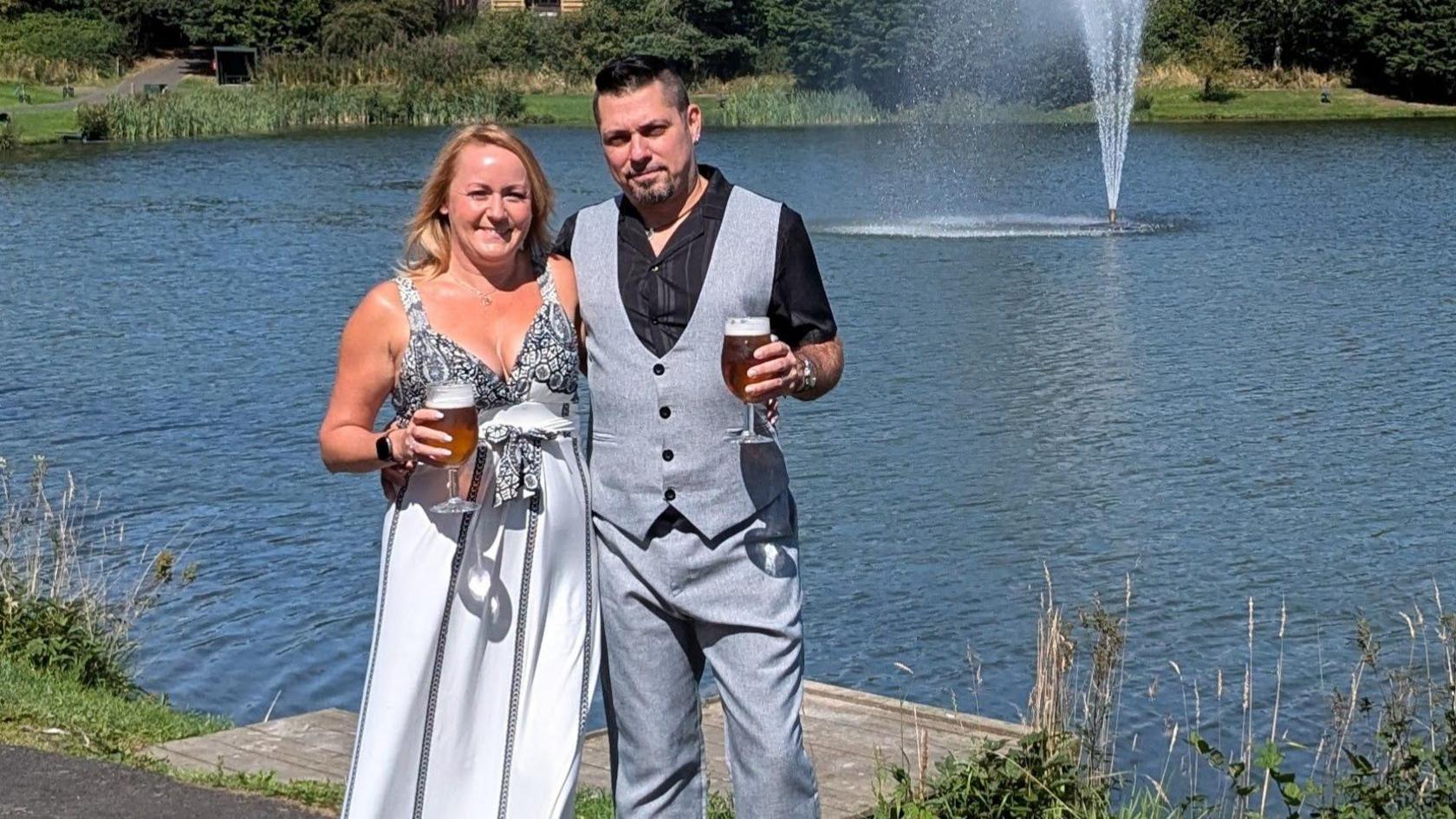 Tracey Watkins and her partner pose in front of a lake. She is wearing a white summer dress and has blonde hair and he is wearing grey trousers and a matching waistcoat. They are both holding a glass of beer.