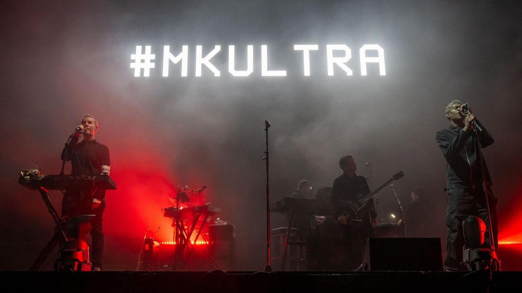 Robert Del Naja and Grant Marshall on stage during the Massive Attack gig on The Downs in Bristol. The screen behind them is glowing red and black and the words "#MKULTRA" are visible in white letters