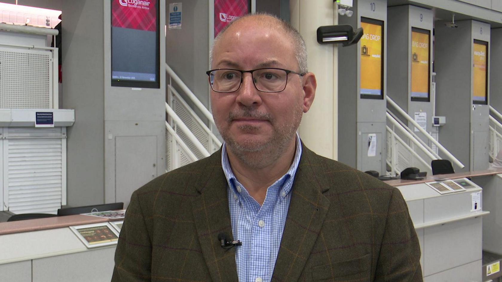A man wearing a suit and shirt standing in front of check-in desks at Jersey Airport.